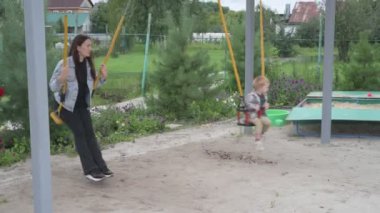 Anne ve küçük oğlu parkta yürüyor, kum havuzunda oynuyor ve salıncakta sallanıyor.