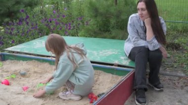 Anne ve küçük kız parkta yürüyor, kum havuzunda oynuyor ve salıncakta sallanıyor.