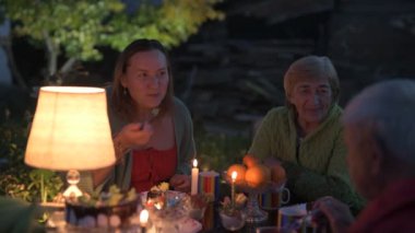 Mutlu yaşlı çift ve yetişkin kızları tatillerini evin avlusunda kutluyorlar, üç kişilik aile lamba ışığında yemek yiyorlar.