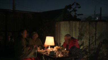 Mutlu yaşlı çift ve yetişkin kızları tatillerini evin avlusunda kutluyorlar, üç kişilik aile lamba ışığında yemek yiyorlar.