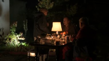 Mutlu yaşlı çift ve yetişkin kızları tatillerini evin avlusunda kutluyorlar, üç kişilik aile lamba ışığında yemek yiyorlar.