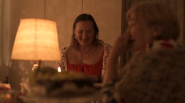 Mutlu yaşlı çift ve yetişkin kızları tatillerini masada kutluyorlar, üç kişilik aile mutfakta lamba ışığında yemek yiyorlar.