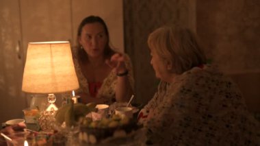 Mutlu yaşlı çift ve yetişkin kızları tatillerini masada kutluyorlar, üç kişilik aile mutfakta lamba ışığında yemek yiyorlar.