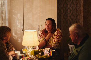 Mutlu yaşlı çift ve yetişkin kızları tatillerini masada kutluyorlar, üç kişilik aile mutfakta lamba ışığında yemek yiyorlar.