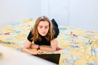 Genç öğrenci kız laptopla çalışıyor, daktilo ediyor ve yataktaki bilgisayar ekranına bakıyor.