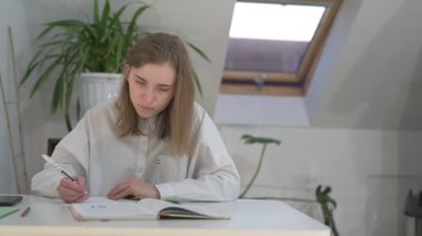 Beyaz tişörtlü liseli kız elinde not defterleri, ders kitapları, akıllı telefon ve dizüstü bilgisayarla masada oturuyor, ödev yapıyor, internetten öğrenim görüyor.