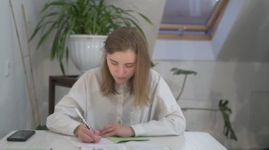 Beyaz tişörtlü liseli kız elinde not defterleri, ders kitapları, akıllı telefon ve dizüstü bilgisayarla masada oturuyor, ödev yapıyor, internetten öğrenim görüyor.