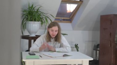 Beyaz tişörtlü liseli kız elinde not defterleri, ders kitapları, akıllı telefon ve dizüstü bilgisayarla masada oturuyor, ödev yapıyor, internetten öğrenim görüyor.