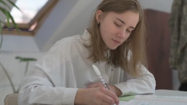 Beyaz tişörtlü liseli kız elinde not defterleri, ders kitapları, akıllı telefon ve dizüstü bilgisayarla masada oturuyor, ödev yapıyor, internetten öğrenim görüyor.