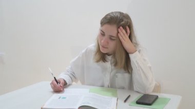 Beyaz tişörtlü liseli kız elinde not defterleri, ders kitapları, akıllı telefon ve dizüstü bilgisayarla masada oturuyor, ödev yapıyor, internetten öğrenim görüyor.