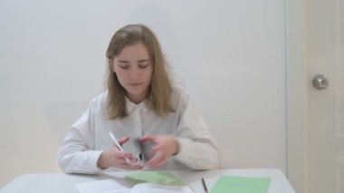 Beyaz tişörtlü liseli kız elinde not defterleri, ders kitapları, akıllı telefon ve dizüstü bilgisayarla masada oturuyor, ödev yapıyor, internetten öğrenim görüyor.