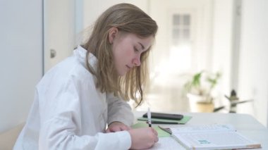 Beyaz tişörtlü liseli kız elinde not defterleri, ders kitapları, akıllı telefon ve dizüstü bilgisayarla masada oturuyor, ödev yapıyor, internetten öğrenim görüyor.