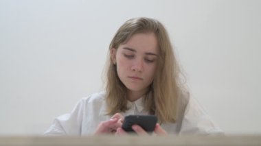 Beyaz tişörtlü liseli kız elinde not defterleri, ders kitapları, akıllı telefon ve dizüstü bilgisayarla masada oturuyor, ödev yapıyor, internetten öğrenim görüyor.