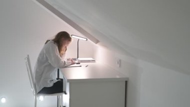 Beyaz tişörtlü liseli kız elinde not defterleri, ders kitapları, akıllı telefon ve dizüstü bilgisayarla masada oturuyor, ödev yapıyor, internetten öğrenim görüyor.