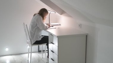 Beyaz tişörtlü liseli kız elinde not defterleri, ders kitapları, akıllı telefon ve dizüstü bilgisayarla masada oturuyor, ödev yapıyor, internetten öğrenim görüyor.