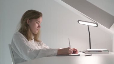 Beyaz tişörtlü liseli kız elinde not defterleri, ders kitapları, akıllı telefon ve dizüstü bilgisayarla masada oturuyor, ödev yapıyor, internetten öğrenim görüyor.