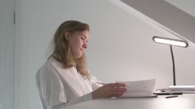 Beyaz tişörtlü liseli kız elinde not defterleri, ders kitapları, akıllı telefon ve dizüstü bilgisayarla masada oturuyor, ödev yapıyor, internetten öğrenim görüyor.