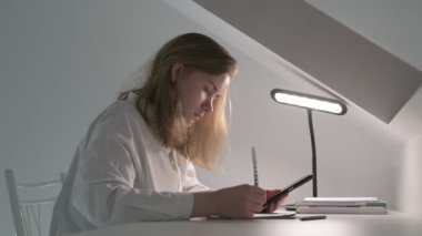 Beyaz tişörtlü liseli kız elinde not defterleri, ders kitapları, akıllı telefon ve dizüstü bilgisayarla masada oturuyor, ödev yapıyor, internetten öğrenim görüyor.