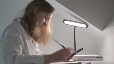 Beyaz tişörtlü liseli kız elinde not defterleri, ders kitapları, akıllı telefon ve dizüstü bilgisayarla masada oturuyor, ödev yapıyor, internetten öğrenim görüyor.