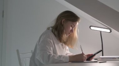 Beyaz tişörtlü liseli kız elinde not defterleri, ders kitapları, akıllı telefon ve dizüstü bilgisayarla masada oturuyor, ödev yapıyor, internetten öğrenim görüyor.