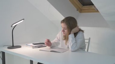 Beyaz tişörtlü liseli kız elinde not defterleri, ders kitapları, akıllı telefon ve dizüstü bilgisayarla masada oturuyor, ödev yapıyor, internetten öğrenim görüyor.