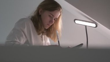 Beyaz tişörtlü liseli kız elinde not defterleri, ders kitapları, akıllı telefon ve dizüstü bilgisayarla masada oturuyor, ödev yapıyor, internetten öğrenim görüyor.