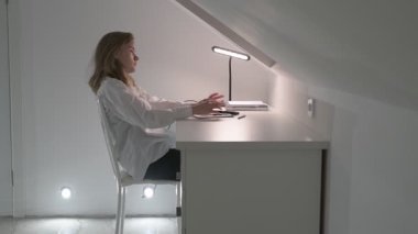 Beyaz tişörtlü liseli kız elinde not defterleri, ders kitapları, akıllı telefon ve dizüstü bilgisayarla masada oturuyor, ödev yapıyor, internetten öğrenim görüyor.