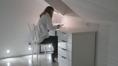 Beyaz tişörtlü liseli kız elinde not defterleri, ders kitapları, akıllı telefon ve dizüstü bilgisayarla masada oturuyor, ödev yapıyor, internetten öğrenim görüyor.