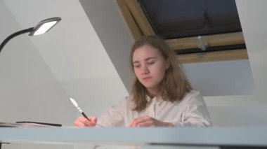 Beyaz tişörtlü liseli kız elinde not defterleri, ders kitapları, akıllı telefon ve dizüstü bilgisayarla masada oturuyor, ödev yapıyor, internetten öğrenim görüyor.