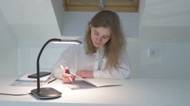 Beyaz tişörtlü liseli kız elinde not defterleri, ders kitapları, akıllı telefon ve dizüstü bilgisayarla masada oturuyor, ödev yapıyor, internetten öğrenim görüyor.