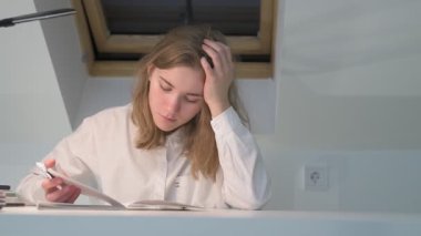 Beyaz tişörtlü liseli kız elinde not defterleri, ders kitapları, akıllı telefon ve dizüstü bilgisayarla masada oturuyor, ödev yapıyor, internetten öğrenim görüyor.