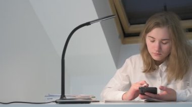 Beyaz tişörtlü liseli kız elinde not defterleri, ders kitapları, akıllı telefon ve dizüstü bilgisayarla masada oturuyor, ödev yapıyor, internetten öğrenim görüyor.