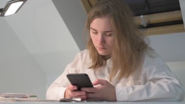 Beyaz tişörtlü liseli kız elinde not defterleri, ders kitapları, akıllı telefon ve dizüstü bilgisayarla masada oturuyor, ödev yapıyor, internetten öğrenim görüyor.