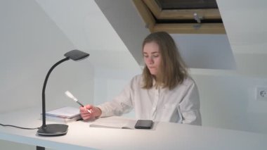 Beyaz tişörtlü liseli kız elinde not defterleri, ders kitapları, akıllı telefon ve dizüstü bilgisayarla masada oturuyor, ödev yapıyor, internetten öğrenim görüyor.