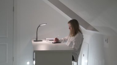 Beyaz tişörtlü liseli kız elinde not defterleri, ders kitapları, akıllı telefon ve dizüstü bilgisayarla masada oturuyor, ödev yapıyor, internetten öğrenim görüyor.