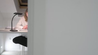 Beyaz tişörtlü liseli kız elinde not defterleri, ders kitapları, akıllı telefon ve dizüstü bilgisayarla masada oturuyor, ödev yapıyor, internetten öğrenim görüyor.