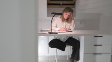 Beyaz tişörtlü liseli kız elinde not defterleri, ders kitapları, akıllı telefon ve dizüstü bilgisayarla masada oturuyor, ödev yapıyor, internetten öğrenim görüyor.