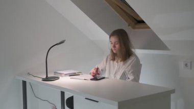 Beyaz tişörtlü liseli kız elinde not defterleri, ders kitapları, akıllı telefon ve dizüstü bilgisayarla masada oturuyor, ödev yapıyor, internetten öğrenim görüyor.