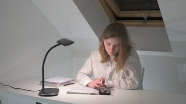 Beyaz tişörtlü liseli kız elinde not defterleri, ders kitapları, akıllı telefon ve dizüstü bilgisayarla masada oturuyor, ödev yapıyor, internetten öğrenim görüyor.