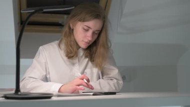 Beyaz tişörtlü liseli kız elinde not defterleri, ders kitapları, akıllı telefon ve dizüstü bilgisayarla masada oturuyor, ödev yapıyor, internetten öğrenim görüyor.