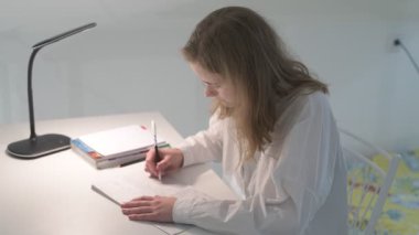 Beyaz tişörtlü liseli kız elinde not defterleri, ders kitapları, akıllı telefon ve dizüstü bilgisayarla masada oturuyor, ödev yapıyor, internetten öğrenim görüyor.