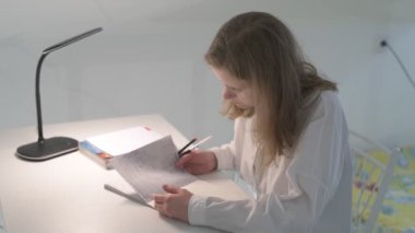 Beyaz tişörtlü liseli kız elinde not defterleri, ders kitapları, akıllı telefon ve dizüstü bilgisayarla masada oturuyor, ödev yapıyor, internetten öğrenim görüyor.