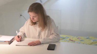 Beyaz tişörtlü liseli kız elinde not defterleri, ders kitapları, akıllı telefon ve dizüstü bilgisayarla masada oturuyor, ödev yapıyor, internetten öğrenim görüyor.