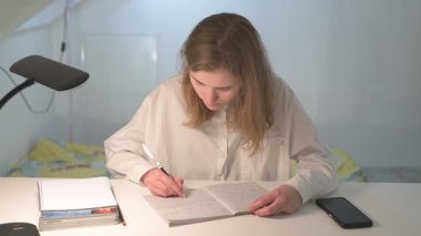 Beyaz tişörtlü liseli kız elinde not defterleri, ders kitapları, akıllı telefon ve dizüstü bilgisayarla masada oturuyor, ödev yapıyor, internetten öğrenim görüyor.