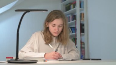 Beyaz tişörtlü liseli kız elinde not defterleri, ders kitapları, akıllı telefon ve dizüstü bilgisayarla masada oturuyor, ödev yapıyor, internetten öğrenim görüyor.