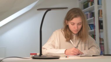 Beyaz tişörtlü liseli kız elinde not defterleri, ders kitapları, akıllı telefon ve dizüstü bilgisayarla masada oturuyor, ödev yapıyor, internetten öğrenim görüyor.