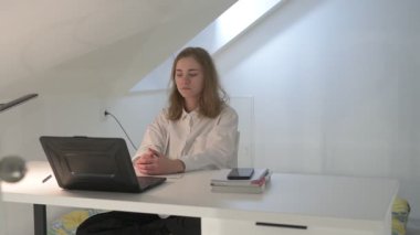 Beyaz tişörtlü liseli kız elinde not defterleri, ders kitapları, akıllı telefon ve dizüstü bilgisayarla masada oturuyor, ödev yapıyor, internetten öğrenim görüyor.