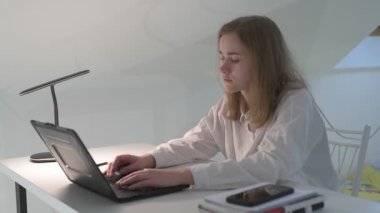 Beyaz tişörtlü liseli kız elinde not defterleri, ders kitapları, akıllı telefon ve dizüstü bilgisayarla masada oturuyor, ödev yapıyor, internetten öğrenim görüyor.