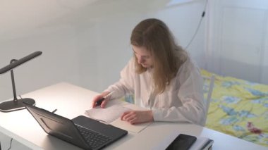 Beyaz tişörtlü liseli kız elinde not defterleri, ders kitapları, akıllı telefon ve dizüstü bilgisayarla masada oturuyor, ödev yapıyor, internetten öğrenim görüyor.