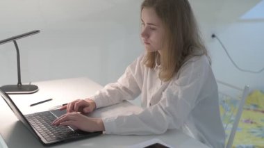 Beyaz tişörtlü liseli kız elinde not defterleri, ders kitapları, akıllı telefon ve dizüstü bilgisayarla masada oturuyor, ödev yapıyor, internetten öğrenim görüyor.